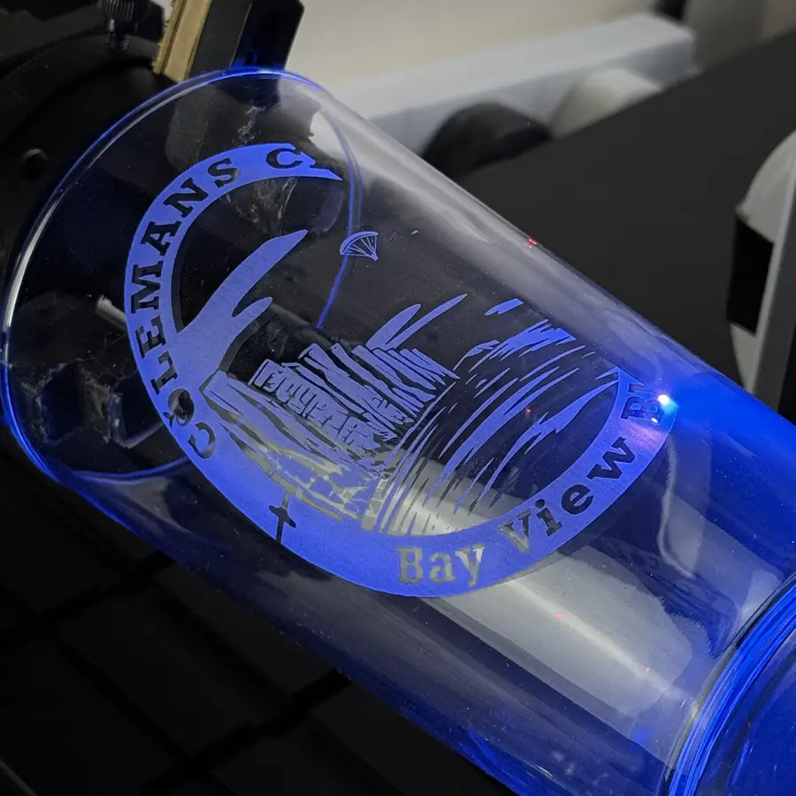 A pint glass on the laser mid-order, lit violet by the UV beam as a crest is engraved
